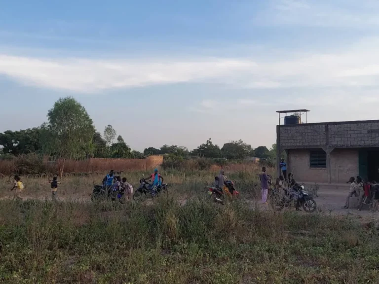 Terrain herbeux au premier plan ; au loin, élèves et motos près d’une classe.
